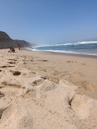 The image depicts a serene beach scene in São João das Lampas, Portugal. The foreground is dominated by sand dunes and footprints, with the ocean and its waves stretching towards the horizon in the background. The water is a vibrant blue, with white-capped waves rolling onto the shore. The sky is a clear, cloudless blue, hinting at a sunny day.
In the mid-ground, a few individuals are enjoying the beach. Two people, a man and a woman, are visible on the sand. The man is kneeling, appearing to interact with something on the ground, while the woman stands a short distance away, facing the ocean. Further down the beach, more people are scattered, enjoying the sun and sea.
To the left of the frame, rugged cliffs rise, adding a dramatic backdrop to the coastal landscape. The cliffs are steep and rocky, with patches of greenery clinging to their slopes. The overall impression is one of a tranquil and picturesque beach, a popular spot for relaxation and recreation.
