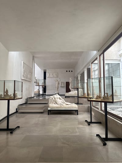 The image depicts the interior of a museum exhibit hall in Canicena, Italy, showcasing classical sculptures. The hall is spacious, with high ceilings and natural light filtering in from large windows on the right. The floor is tiled with large, pale grey stone slabs.
Dominating the foreground is a headless marble sculpture of a reclining nude figure. To the left, a similar marble sculpture of a nude figure is displayed on a raised platform. In the middle ground, a set of grey stone steps leads up to a slightly elevated area where more sculptures are displayed. One prominent sculpture on this level is a standing, draped female figure.
Several glass display cases on black metal stands are positioned throughout the hall, containing smaller sculptures and artifacts. These cases are illuminated, drawing attention to their contents. In the background, more relief sculptures are mounted on the white walls, and various statues are visible. The overall impression is one of an organized and well-lit exhibition space dedicated to classical art. The environment appears clean and well-maintained. No people are visible in the image.