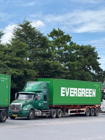 This media file shows a large green semi-truck parked on a paved surface, possibly a roadside or a parking lot. The truck is carrying a bright green shipping container that has "EVERGREEN" printed in large white letters on its side. Another green shipping container is partially visible at the bottom of the frame. The truck's cab is also green and displays markings that include "MECS" and "HOANG GROUP" along with numbers like "3H-117.35" and "43H-117.35". In the background, there are lush green trees and a bright blue sky with scattered white clouds. Power lines are also visible stretching across the sky. The overall scene suggests a sunny day in a location with natural vegetation, typical of a place like Lao Bảo Commune, Vietnam, where such logistics operations would occur. There are no people visible in the frame.
