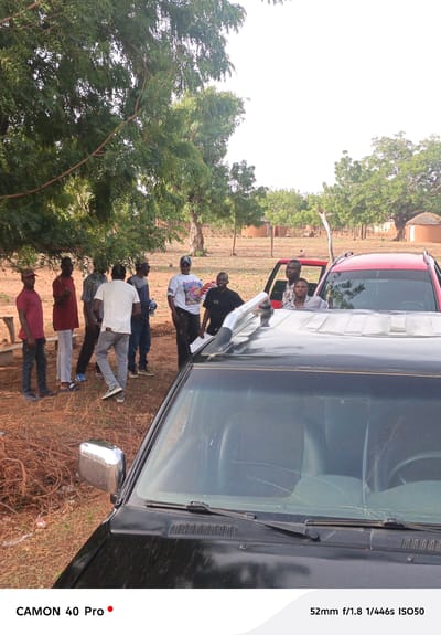 In Gidan Kogi, Nigeria, a group of approximately ten men stand and interact near two vehicles, one red and one black, under a tree. In the background are a few huts typical of the area. The foreground is dominated by the black vehicle, showing the hood, windshield with wipers, and a side mirror. The red vehicle is partially visible behind it. The men appear to be engaged in conversation. The environment is dry, with reddish-brown soil and scattered dry branches on the ground.