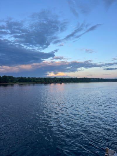 The image depicts a serene waterfront scene at dusk in Nevskaya Dubrovka, Russia. A vast expanse of calm, dark blue water occupies the foreground, with gentle ripples reflecting the muted colors of the sky. In the middle ground, a dense line of dark green trees forms the shoreline, suggesting a forested area. The sky above is a gradient of deep blue, transitioning to lighter shades near the horizon. Scattered clouds, some dark and ominous, others wispy and light, are present throughout the sky. A faint glow of orange and pink hues is visible on the horizon, indicating the setting sun. The overall atmosphere is tranquil and natural, characteristic of a quiet evening by the water. There are no people or specific man-made structures prominently visible, other than a small portion of a white railing or structure in the bottom right corner, possibly part of a boat or dock.