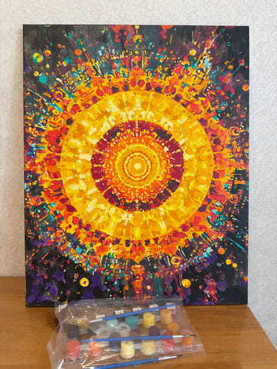 The image presents a close-up, high-angle view of a completed paint-by-numbers project depicting a vibrant, abstract circular design reminiscent of a sun or explosion. The artwork is on a canvas, predominantly featuring concentric rings of warm colors including yellow, orange, and red, set against a darker background filled with multicolored flecks and streaks. The canvas sits on a light-colored surface, possibly a tabletop. To the right of the canvas, a clear plastic bag contains multiple small paint pots with corresponding brushes, suggesting the materials used for the paint-by-numbers activity. The details present no indication of Myaglovo, Russia.