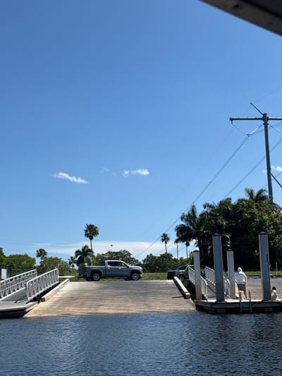 The image depicts a sunny day at a boat ramp in Fort Lauderdale, United States. The scene includes a concrete ramp leading into the water, where ripples suggest slight movement. A gray pickup truck is parked on the ramp. Palm trees and green vegetation line the side of the ramp, contrasting with the clear blue sky marked by a few wispy clouds. Electrical power lines stretch overhead. A dock extends into the water on the right. One or two people are on the dock. Metal poles are on the dock.