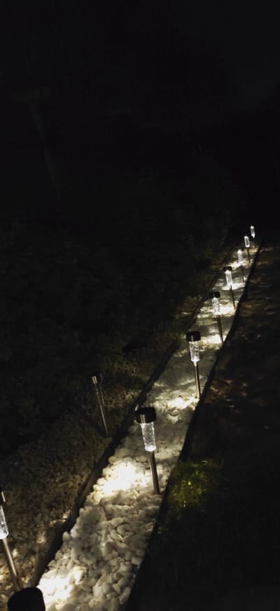 A ground-level, vertical, outdoor shot at night depicts a path lined with solar-powered garden lights. The path is comprised of a narrow channel filled with white gravel, bordered by darker soil or mulch. A row of clear, cylindrical solar lights with black tops are spaced along the gravel path, emitting a warm, white glow. These lights illuminate the gravel and cast light onto the surrounding dark ground. In the upper left portion of the image, the ground appears to be covered in darker, more textured material, possibly soil or mulch with some low-lying vegetation. The overall scene suggests a well-maintained garden or pathway in a residential or park setting. The image does not contain any specific landmarks that would identify it as being located in the city of Pleszew, Poland, or any other specific geographical location.