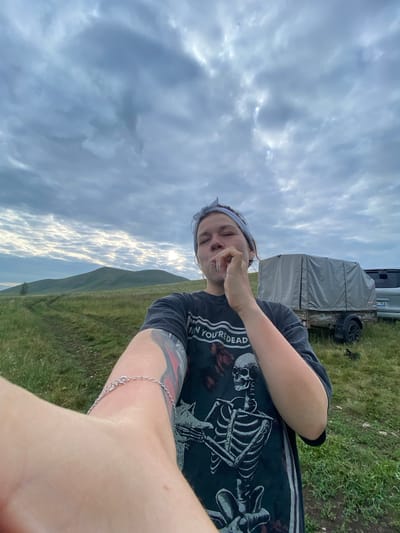 In a grassy field under a cloudy sky, a young woman is taking a selfie. She is wearing a dark t-shirt with a skeleton graphic and the text "WHEN YOU'RE DEAD". A light-colored bandana is tied around her head. Her right hand is raised to her mouth, holding a white object, possibly a cigarette or vape pen. A silver bracelet is visible on her left wrist, which is extended towards the camera. In the background, a gray-covered trailer and a portion of a silver vehicle are parked on the grass. Rolling hills are visible in the distance, with a single tree on the left side of the frame. The location is identified as the village of Абзаново, Russia.