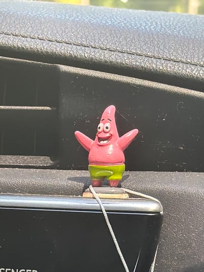 A pink figurine of Patrick Star from SpongeBob SquarePants, wearing green shorts, is affixed to the dashboard of a vehicle. The figurine is positioned in front of a black air vent and rests on a silver trim piece. A white string is attached to the figurine and dangles down. The background shows the textured black interior of the car, with a hint of sunlight and greenery visible through the windshield. The image was taken in Seaside, United States.