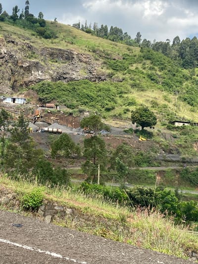 The image presents a panoramic, elevated view of a hilly, green landscape, indicative of Kinyona ward, Kenya. The scene is largely dominated by what appears to be a quarry or a substantial construction site carved into the side of a steep, rocky hillside.
In the central and lower portions of the image, the active site is bustling with heavy machinery and materials. Prominent yellow conveyor belt systems crisscross the area, leading to and from what look like crushing or sorting units. A yellow excavator is visible, along with multiple trucks; one carries several large cylindrical tanks, while another appears to be a dump truck. Piles of gray aggregate, likely crushed rock or gravel, are scattered across the excavated, disturbed earth.
The surrounding natural environment features a mosaic of green vegetation. Rocky outcrops and cliff faces are interspersed with areas of dense trees and grassy slopes. A thick forest canopy crowns the upper part of the hill against an overcast, cloudy sky, suggesting a daytime setting, possibly late morning or afternoon.
To the right of the frame, a paved asphalt road with a white dividing line indicates a higher vantage point. Beyond this road, more lush greenery, including trees and undergrowth, extends into the distance. Power lines and utility poles are visible across the landscape, denoting existing infrastructure. A single individual can be faintly seen walking on a path or smaller road in the upper right section of the image. No discernible text is visible.