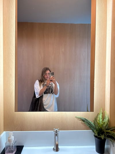 A person is reflected in a large, rectangular mirror with a wooden frame and soft, built-in lighting. The person is holding a smartphone and taking a picture of themself. The reflection shows a white t-shirt, glasses, and long brown hair. To the right of the mirror is a white sink with a metal faucet and a green fern in a black pot sitting on the countertop. Below, partially visible, is a soap dispenser. The background appears to be a wood-paneled wall, also seen in the mirror's reflection.