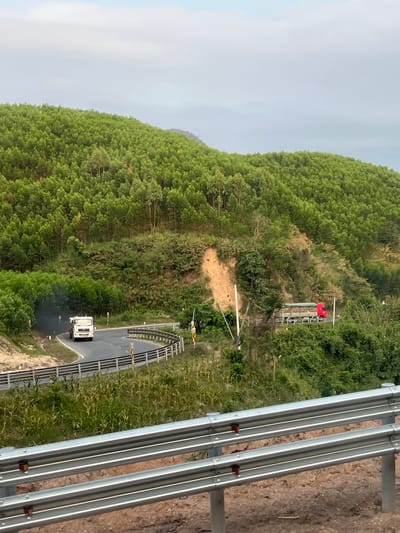 The image depicts a hilly landscape in Aê Thi, Vietnam, densely covered with green trees and foliage. A winding road cuts through the landscape, with a white truck visible on the road. To the right of the road is a metallic guardrail, and further up the hill, there are structures that might be part of some kind of communication tower, distinguished by a red flag on top. The sky is overcast and pale.