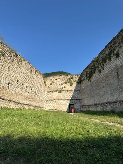 The image shows a view of the Khuchni fortress in Russia under a clear blue sky. The fortress walls are made of stone and are overgrown with vegetation in places. A person wearing a red jacket is seen standing in a doorway in the distance, within the fortress walls. The foreground is covered in green grass, with shadows cast by the walls.