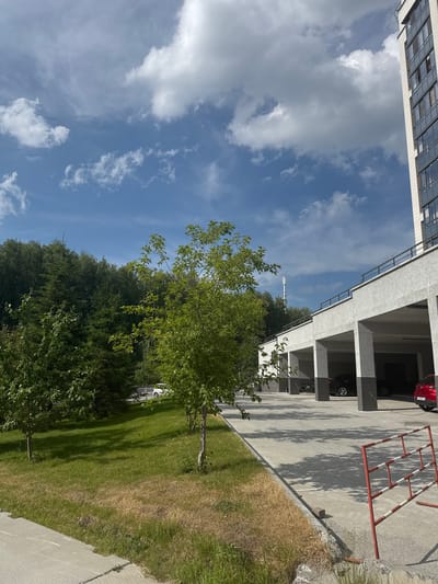 The image depicts an outdoor scene in Koltsovo, Russia, featuring a modern apartment building with a covered parking area. The parking area has multiple concrete pillars supporting an upper level and a black railing along the edge. Several cars are parked within the covered bays, including a black sedan and a red hatchback. In the foreground, a patch of grass is visible, with some areas appearing dry and brown, indicating dry conditions. A red and white striped barrier is positioned in the lower right corner of the image. To the left of the parking structure, a line of trees and dense green foliage forms a backdrop. The sky above is a clear blue with scattered white clouds. A tall, slender telecommunications tower is visible in the distance, rising above the trees. The scene appears to be a residential area, possibly a courtyard or common space within a housing complex.