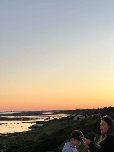 The image depicts a sunset view from a location overlooking a body of water in Vila Real de Santo António, Portugal. A woman and a boy are positioned on the right side of the frame, observing the scene. The woman is holding a phone near the boy's head, possibly taking a picture or showing him something on the screen. The backdrop features a blend of natural elements and man-made structures. There is a coastline densely covered with vegetation and houses, a mix of wetlands, channels, and water surfaces, and the sky during sunset is a gradient from light blue to orange.