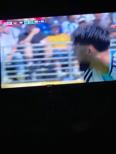The image displays a close-up of a television screen showing a football match. In the upper portion of the screen, a scoreboard indicates the time as 01:25, with the score at ESP 0 - 0 ARG. The flags of Spain (ESP) and Argentina (ARG) are visible next to their respective scores. The background of the screen shows blurred images of spectators in a stadium. A player, likely from the Argentinian team due to the kit colors, is partially visible on the right side of the screen, facing away from the camera. The bottom portion of the image is dark, suggesting the room is dimly lit, and a small red indicator light is visible on the television bezel. There is no indication or visual information within the image that places this scene in the city of Legenda, Egypt.