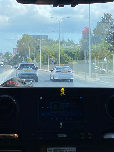 The image displays a daytime street view from the interior of a vehicle in Nahariya, Israel. The foreground reveals a dark dashboard with an infotainment screen, USB ports, and a yellow ribbon affixed above the screen. A black and red gloved hand is visible on the lower left of the windshield. A sticker with partially legible Hebrew text, indicating a safety monitoring system for drivers and passengers, is adhered to the dashboard.
The perspective is forward onto a multi-lane road with a median. Immediately ahead, a light-colored SUV with a yellow Israeli license plate is positioned in front of a white sedan. The sedan, also displaying a yellow Israeli license plate and a yellow roof sign, is operating as a taxi. Both vehicles appear stationary. A red traffic light is visible in the distance on the left. The environment includes green trees, palm trees, and a tree with pink blossoms lining the road. Streetlights are present, and in the far background, a building with a multi-tiered roofline and a construction crane are faintly visible above the treeline.