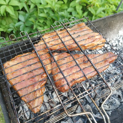 The image shows three pieces of marinated meat cooking on a metal grill over hot coals. The grill is positioned in a barbecue pit made of concrete. In the background, lush green foliage is visible. The scene appears to be an outdoor setting, likely a backyard or park, in the city of Luzino, Russia. The meat is glistening with marinade and has grill marks from the hot bars. The coals are glowing and appear to be providing consistent heat for cooking. The overall impression is one of outdoor cooking and leisure.