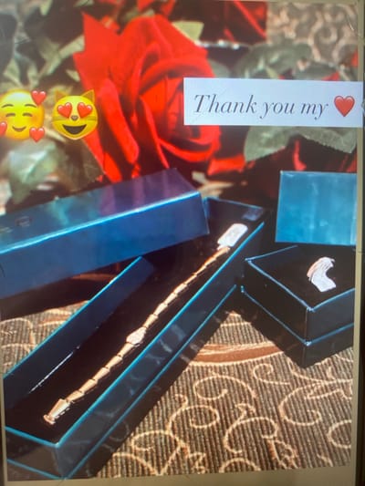 This image depicts a close-up view of a gift presentation, likely a jewelry item, set against a backdrop of red roses. In the foreground, there are two open gift boxes, one dark blue and the other black. Inside the dark blue box, a delicate, segmented bracelet or necklace is displayed on a black velvet lining. The black box contains a single, light-colored, intricately designed ring.
A white rectangular sticker overlaying the roses features text in a cursive font that reads, "Thank you my" followed by a red heart emoji. To the left of the text, two emoji faces are superimposed on the image: one with closed eyes and rosy cheeks, and another cat face with heart-shaped eyes. The background is dominated by large, vibrant red roses with green foliage, suggesting a romantic or celebratory occasion. The overall impression is one of gratitude and a thoughtful gift.
There is no visible indication of the city of Arbagish, Uzbekistan, or any specific urban environment. The scene is focused on the gifts and the accompanying message.