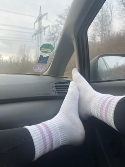 The image displays an interior perspective from the passenger side of a vehicle, looking forward and slightly right, consistent with an environment in Rheinfelden (Baden), Germany. Two feet, clad in white ankle-length socks with three horizontal lavender stripes and black leggings, are elevated and rest with their heels on the lower section of the grey dashboard, positioning the soles towards the camera. The dashboard features an "SRS AIRBAG" label and an air vent. A slender green stem or stick lies horizontally on the dashboard beneath the windshield. Affixed to the windshield are two German environmental stickers: a prominent green circular "UMWELT ZONE 4" sticker and a smaller, partially obscured purple circular sticker beneath it. Through the windshield, under an overcast sky, a large metallic electricity pylon supporting power lines is visible in the middle ground, with additional pylons or sections of the structure further in the background. A sparse forest of bare deciduous trees stretches across the landscape, indicating a winter or late autumn season. The exterior side mirror on the passenger side reflects the sky and trees.
