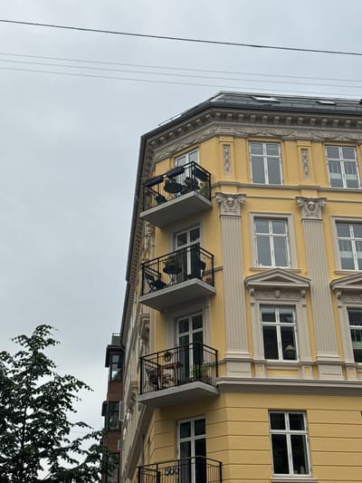 The image shows the exterior of a building on a cloudy day. The building is yellow with white trim and several balconies with black railings. There are trees to the left of the building. The image appears to be taken from a street level perspective, looking up at the building. There are no people visible in the image. The building has multiple windows, some of which appear to be on different floors. There are also some decorative architectural elements on the building. The sky is overcast and gray, and there are power lines visible in the upper portion of the image.