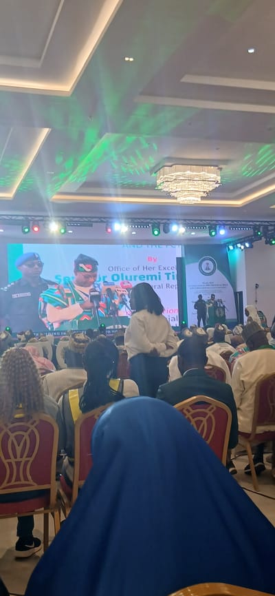 The image shows an indoor event in a large room, likely in Unguwan Sanusi, Nigeria, based on the onscreen text about Oluremi Tinubu. There is a large screen on stage displaying images and text. Several people are seated in chairs, facing the stage. A person stands at a podium on the screen, likely giving a presentation. The room has modern lighting, including a large chandelier, and green lighting effects on the ceiling.