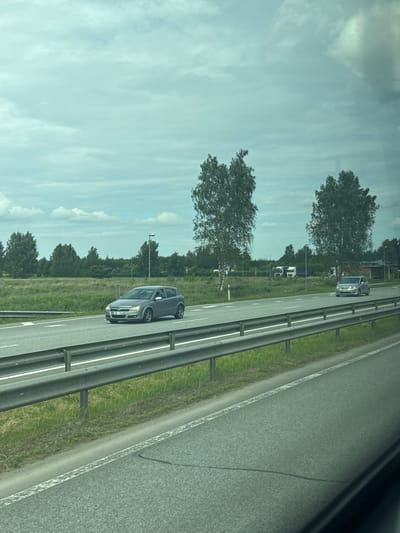 This media file shows a view from a vehicle traveling along a highway in Jaunolaine, Latvia. The scene is dominated by a two-lane road with a metal guardrail separating it from the grassy shoulder. The road appears to be a highway, indicated by the smooth asphalt and lane markings.
Several cars are visible on the road, including a dark gray car in the foreground on the right lane. Further down the road, another dark gray car is visible in the left lane. In the distance, a white truck with a blue cab is parked on the side of the road, possibly at a rest stop or service area.
The landscape flanking the highway is green and lush, with numerous trees lining the background. The sky is partly cloudy with a light blue hue, suggesting it is daytime, likely mid-morning or afternoon, with fair weather. The overall impression is of a typical journey on a highway in a rural or suburban setting. There is no visible text in the image.