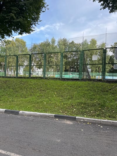 A paved road borders a grassy embankment topped by a green chain-link fence. Beyond the fence, a tennis court with blue and green surfaces is visible, along with a white building and trees. The sky is partly cloudy. The location is identified as Putyatino (Semivragovskaya S/A), Russia.