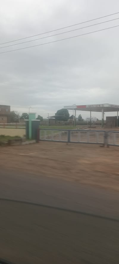 The image, taken from a moving vehicle, shows the exterior of a filling station in Ladura Foron, Nigeria. The station features a covered refueling area with visible signage, likely indicating the brand and services offered. A large white cylindrical tank, presumably for fuel storage, is visible behind the main structure. The foreground includes a gray metal fence, a short wall, and an expanse of brown, unpaved ground. Overcast sky is visible above. Utility lines run across the top portion of the frame. A building with square windows is to the left of the frame. The image has motion blur.