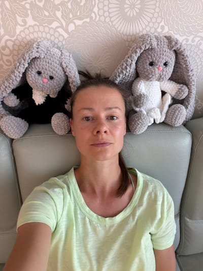 A woman is sitting on a light gray couch, leaning against a patterned wall. To her left and right are two large, crocheted gray bunnies. The bunny on the left is wearing a black outfit with a white collar. The bunny on the right is wearing a white outfit. The woman has medium-length brown hair pulled back and is wearing a light green t-shirt. The scene appears to be indoors, possibly in a home in Melick, Netherlands, given the wallpaper and couch. The lighting suggests it is daytime.