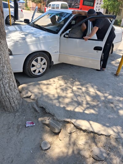 In Kahramon, Uzbekistan, on a sunny day, a white sedan with its driver's side door open is parked on a cracked concrete surface. A man wearing a black t-shirt, black pants, and a dark beanie stands leaning into the open driver's door, talking to the driver. The driver, wearing a light-colored shirt, is visible behind the steering wheel. In the foreground, near a tree trunk, there is scattered trash, including cigarette butts and a crumpled red and white wrapper. In the background, a red bus and a white van are parked, suggesting a street or parking area within a city. The ground is uneven with broken concrete and scattered stones.