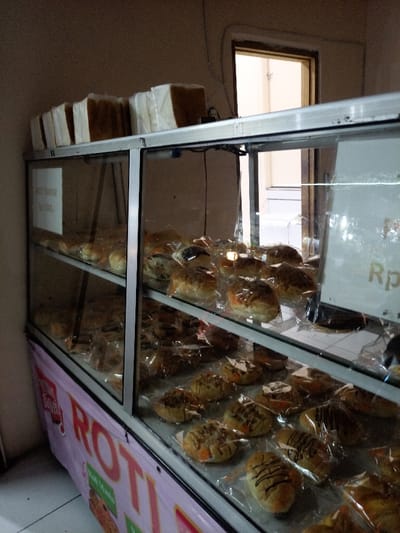 The scene depicts the interior of a retail space, specifically a bakery or bread display area, located in the city of Caringin, Indonesia. A prominent two-tiered glass display case with an aluminum frame is positioned against a plain, light brown wall. On the top surface of this display case, approximately ten rectangular loaves of bread, individually wrapped in white paper or plastic, are stacked horizontally.
The glass shelves inside the display case are filled with numerous individually packaged baked goods. The items are primarily sweet buns or bread rolls, each encased in clear plastic wrap, displaying various toppings such as chocolate drizzle and sprinkles. A white sign affixed inside the upper left section of the display case reads "ROTI TAWAR" and "Rp 7.000,-". A light-colored banner, partially visible and attached to the lower front of the display case, features the word "ROTI" in large red lettering, with smaller text "Roti Manis" also discernible.
In the background, an open doorway reveals an adjacent interior space characterized by brighter illumination, white or very light-colored walls, and white tiled flooring. The overall lighting within the primary scene appears artificial and somewhat dim. No individuals are present in the image, and no actions or interactions are depicted.