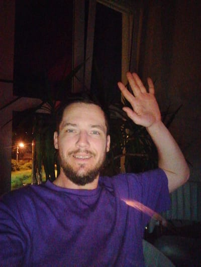 The image shows a close-up, selfie-style shot of a light-skinned man with a beard and blue eyes, wearing a purple shirt. He is smiling and waving with his right hand, which is raised. The background is dark, with some blurry green foliage and a light visible through a window. The lighting suggests an indoor setting at night, with a street lamp visible outside. The image quality is grainy, possibly due to low light conditions. The exact location of Trubacheyevka, Russia, is not discernible from the visual cues in the image.