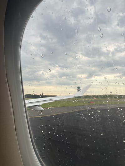 The image is an exterior view from an airplane window during a rain shower, looking out onto an airport tarmac. Water droplets are visible on the window. The airplane's wing is prominent in the foreground, extending across the frame. In the middle ground, a runway or taxiway is visible, with green grass and trees beyond. A control tower with a distinctive structure is situated in the distance. The sky is overcast with dark clouds. The location is identified as Steenokkerzeel, Belgium, suggesting this is likely Brussels Airport. There are no people or interactions depicted in the image. The scene is static and appears to be pre-takeoff or post-landing.