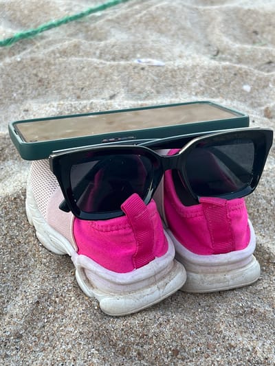 The image shows a pair of pink and white running shoes, a pair of black sunglasses, and a phone resting on sand. The sunglasses are perched on top of the shoes, and the phone is leaning against the sunglasses. The location is likely Kalmunai, Sri Lanka, based on geographical associations. The scene depicts personal belongings placed on a sandy surface.