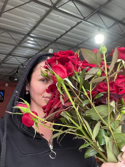 A woman wearing a black hooded jacket and a gold earring is partially obscured by a bouquet of red roses. She has dark hair and is looking towards the camera with a slight smile. The background appears to be an indoor space with a corrugated metal ceiling and exposed metal beams. A bright light fixture is visible hanging from the ceiling. The scene is likely indoors, possibly a market or a covered outdoor area, in Dubăsari town council, Moldova.