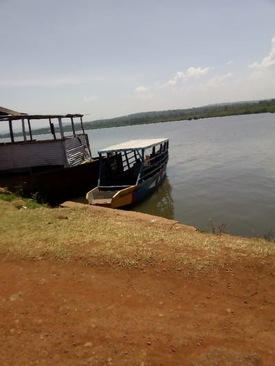 The image displays a daytime scene at a waterfront location in Kendu Bay, Kenya. In the foreground, reddish-brown earth is visible, featuring sparse patches of dry, light green grass and small stones, sloping towards the water's edge.
In the midground, two wooden vessels are docked. The boat on the left is larger and appears to be in a state of disrepair, with a dark hull and a raised, open-sided structure partially covered with corrugated metal sheets. To its right, a smaller passenger boat is moored. This vessel has a blue hull with yellow and red trim, and is fitted with a white canopy over its open seating area. Several indistinct human figures are seated within this passenger boat. Yellow lettering, likely "WEED KOKO", is visible on the red-painted stern of this boat. The water is dark and calm, showing reflections from the sky.
The background features a distant, undulating landmass densely covered with green vegetation, extending across the horizon. The sky is light blue with scattered white cumulus clouds, indicating bright daylight conditions.