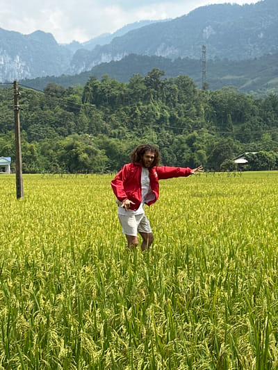 An outdoor, brightly lit, upright portrait-oriented image taken in Ngọc Đường Commune, Vietnam, captures a person posing in a lush rice field. The individual, appearing to be an adult male with long, dark, curly hair, is positioned slightly right of the center of the frame. He is wearing a bright red long-sleeved jacket over a white top and light-colored shorts. His body is turned slightly to the left, and he is looking directly at the camera with a joyful or playful expression. His left arm is raised, with his hand forming a "peace" or "V" sign, while his right arm is bent, and his hand is near his waist. He appears to be barefoot.
The foreground and midground are dominated by a vibrant green rice field, with individual rice plants visible, some appearing to be ripening to a light yellow hue. The field extends into the distance, meeting a line of dense, dark green trees and forested mountains. In the background, layers of mountains recede into a lighter blue and grey haze, suggesting distance and depth, under a light blue sky with some soft white clouds.
To the left of the frame, a wooden utility pole stands, and power lines stretch across the mid-ground. Further back, a metal lattice structure, possibly a transmission tower, is faintly visible against the distant mountains. The overall impression is one of natural beauty and an active, happy moment in an agricultural landscape. There is no visible text in the image. The time of day appears to be mid-morning or afternoon, given the bright, direct sunlight.