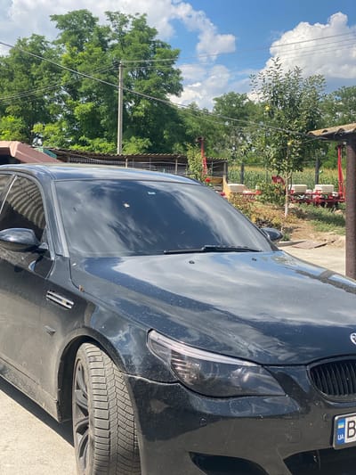 The image shows a black BMW sedan parked outdoors, likely in a rural or semi-rural setting given the background of trees, a field, and what appears to be agricultural equipment. The car's windshield is visible, reflecting the cloudy sky. In the background, there are power lines and utility poles, suggesting the presence of infrastructure. Further back, there's a cornfield, some bushes, and what looks like a simple structure or shed. The location is identified as Pletenyi Tashlyk, Ukraine. There are no people visible in the image. The car is parked on a paved or dirt surface.