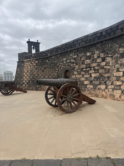 The image depicts cannons at Castillo de San Gabriel, a historic fort in Arrecife, Spain. The foreground features two cannons with wooden wheels resting on a paved stone surface. The cannons point towards the viewer, with a portion of the fort's stone wall rising behind them. The wall is made of dark, weathered blocks and includes a small arched window. In the background, below the fort and cannon, a modern building can be seen, partially obscured. Above the fort, the sky is overcast.