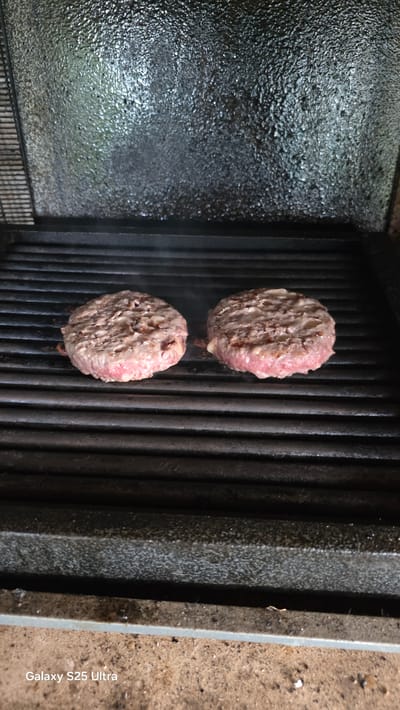 Two raw hamburger patties are being grilled on a black, ridged grill. The patties are round and thick, with visible ground meat texture. Some of the fat is beginning to render and melt, creating small pockets of gooeyness on the surface of the patties. A faint wisp of smoke rises from the rightmost patty. The grill appears to be outdoors, as evidenced by the rough, sandy ground visible in the foreground and the textured, dark metal lid above the grill, which reflects ambient light. The overall lighting suggests it is daytime. The image contains the text "Galaxy S25 Ultra" in the bottom left corner. The scene is likely in Ezerovo, Bulgaria, a small village.