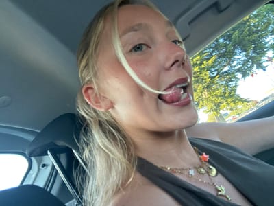 The image presents a playful, close-up portrait of a young woman situated inside a car. She has blonde hair and fair skin, with strands falling across her face. Her blue eyes are cast slightly upwards as she looks towards the camera, sticking out her tongue in a cheerful, lighthearted gesture. Her mouth is open, revealing her tongue and teeth, which appear to have braces. She is wearing a dark, possibly olive green, top or dress and accessorizes with multiple layered gold necklaces featuring various charms, including a red starfish, a seashell, a white flower, and a green bird.
The setting is within a vehicle, with parts of a dark headrest and the grey interior ceiling visible in the frame. Through the car windows, a bright, sunny outdoor environment is apparent, dominated by vibrant green trees. In the background, beyond the trees, several national flags displaying colors like red, yellow, and black are visible on poles, suggesting a European location. Faint outlines of other vehicles can also be seen in the distance, indicating a street or parking area. The strong lighting suggests it is daytime with clear weather. There is no visible text in the image. The overall impression is candid and jovial.