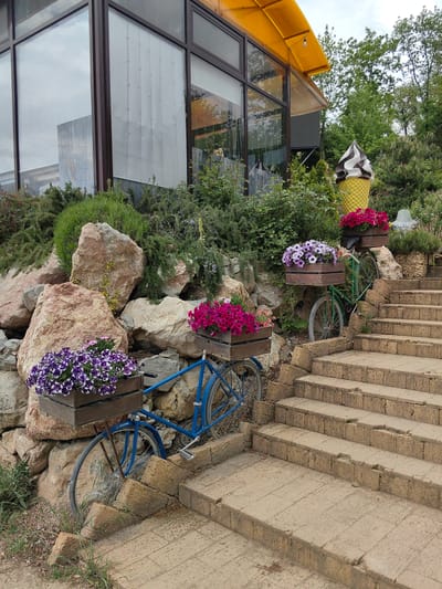 The scene depicts an outdoor display in Luchistoe, Ukraine. Two bicycles, one blue and one green, are positioned beside a set of stone stairs. Each bicycle has a wooden planter box attached, filled with colorful flowers. A large, simulated ice cream cone stands behind the green bicycle. The bicycles and stairs are surrounded by rocks, greenery, and a building with large windows and a yellow awning. The sky appears overcast.
