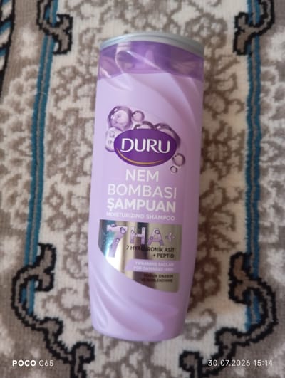 A partially visible, light purple shampoo bottle with a silver cap is placed on a patterned rug. The bottle has branding that includes the word "DURU" in a purple oval and text indicating "NEM BOMBASI ŞAMPUAN" and "MOISTURIZING SHAMPOO". Below this, there is text in a smaller font, and a silver foil label with the imprint "7 HA+" and smaller text that appears to read "7 HYALURONİK ASİT + PEPTID" and "YIPRANMIŞ SAÇLAR FOR DAMAGED HAIR". Further text below says "YOĞUN ONARIM VE NEMLENDİRME". The rug has a repeating pattern of gray, black, and light blue. The bottom left of the image shows "POCO C65" and the bottom right shows a date and time stamp "30.07.2026 15:14". The scene is indoors, and there are no people visible. The city of Beruniy, Uzbekistan is not depicted in this image, nor is there any visual information that would suggest its location.