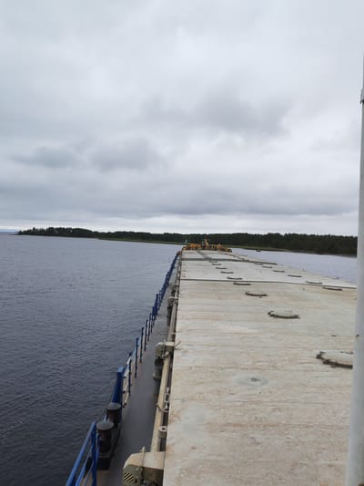 The image shows a view from a barge on a cloudy day, looking towards a forested shoreline and a body of water. The barge is made of concrete and has a series of circular hatches on its deck. A blue railing runs along the side of the barge. In the distance, a white buoy is visible in the water, and further along the shoreline, construction equipment, possibly cranes, can be seen. The water appears calm, and the sky is overcast with grey clouds. The location is identified as Shal'skii, Russia.