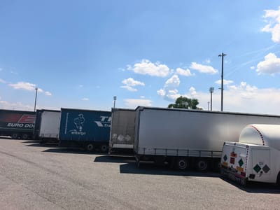 The image depicts a parking lot filled with commercial trucks under a bright, partially cloudy sky. The trucks are parked in a line, and their trailers are predominantly white or blue, with some bearing company logos and text. One trailer is blue with the logo "entargo" and the website "www.transpress.bg". Another green trailer has the logo "EURO DON" printed on it. The parking surface appears to be asphalt. In the background, several tall poles, possibly for lighting or communication towers, are visible, along with some trees on the right side of the frame. The scene is static, showing the trucks at rest. There are no people visible in the image. The location is identified as Sid Municipality, Serbia.