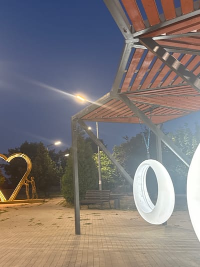 The image captures an outdoor scene at dusk in Egorlykskaya, Russia. In the foreground, a paved walkway leads towards a park area. A large, illuminated heart-shaped sculpture with two human figures is visible on the left. To the right, an abstract, oval-shaped light fixture hangs from a modern, open-sided canopy structure with a wooden slatted roof. In the background, trees and streetlights line the pathway, and a park bench is situated near a cluster of trees. The sky is a deep twilight blue, with a bright street light casting a glare.