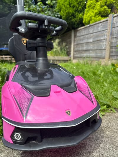 A close-up, eye-level, outdoor shot focuses on the front of a pink toy Lamborghini car. The car, with its black steering wheel and dashboard visible, sits on a concrete path surrounded by green grass and a wooden fence in the background. The car features the Lamborghini logo on its hood and decorative geometric patterns on the front fenders. The scene appears to be in a residential garden, possibly in the city of Oldham, United Kingdom, given the style of the fence and the general appearance of the greenery. The lighting suggests it is daytime with the sun casting shadows. There are no people visible in the image.