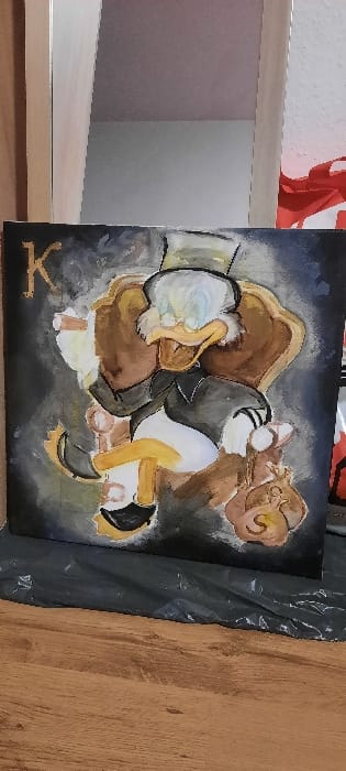 An indoor scene in Kerpen, Germany, depicts a rectangular canvas painting angled towards the viewer, resting on a crumpled black plastic sheet placed directly on a light brown wooden floor. The painting features an anthropomorphic duck character, resembling Scrooge McDuck, centrally positioned. This character, with a yellow bill, white facial hair, and spectacles, is depicted sitting in a dark brown, ornate armchair. He wears a dark suit jacket, a lighter vest or shirt, and a grey top hat. In his raised left hand, he holds a lit cigar. His right hand rests on the armrest of the chair, near a brown money bag with a distinct dollar sign. The background of the painting is predominantly dark, possibly black or very dark grey, with lighter, brush-stroked highlights. A golden letter "K" is visible in the upper left corner of the canvas. Behind the painting, a vertical light-colored wooden or laminate structure is visible on the left, while a white wall and a portion of a red object are visible on the right. A reflection of a room with a light ceiling and a grey wall is present in the upper central background, suggesting a reflective surface behind the artwork.