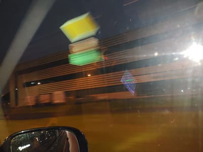 The image captures a view through a car window, likely in Laagri alevik, Estonia, at night or in low light conditions. A blurred reflection of stacked, illuminated objects, possibly signs or lights, appears in the upper left quadrant of the frame. These are colored yellow, orange, and green, and are out of focus due to motion. In the middle ground, a building with horizontal lines, possibly corrugated siding or architectural elements, is visible. The building has a dark upper section and a lighter, segmented lower section. A bright light source, possibly a streetlight or headlights, causes significant glare and lens flare on the right side of the image. In the lower left corner, a side-view mirror of the car is visible, reflecting blurred lights and a white sign with some text, including what appears to be "Darinem". The overall impression is a nighttime drive with abstract reflections and lighting elements.
