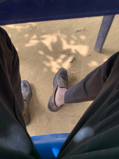 The image provides a close-up, downward-facing perspective of two human legs situated on light brown, unpaved ground in Assai Sabongida, Nigeria. The ground has a sandy or dusty texture and is illuminated by natural light, casting distinct, dappled shadows across its surface, indicative of overhead foliage. Both visible legs are clad in dark brown trousers. The foot on the left is fully encased in a brown leather loafer. The foot on the right features a similar brown leather loafer, but it is partially slipped off the foot, exposing the bare heel and lower ankle. A segment of a blue object, consistent with a chair or bench seat, is visible at the bottom of the frame, positioned between the legs. Additionally, a blue, possibly metallic, furniture leg is present in the upper right corner of the image. The scene appears static.