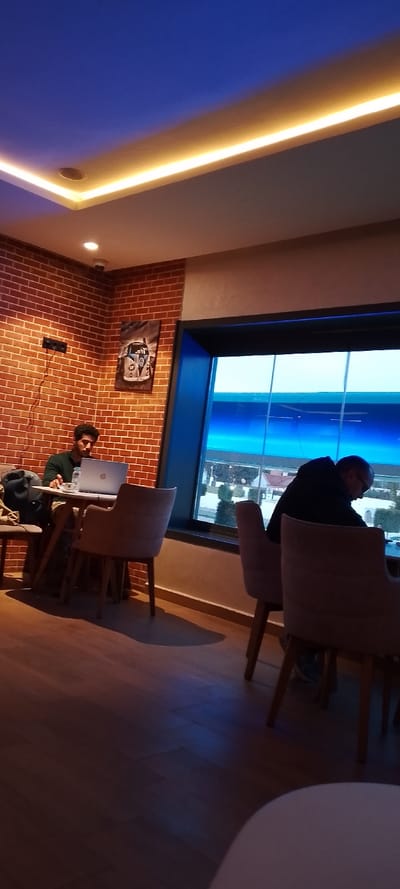 Two men are sitting in a cafe. One man is at a table, working on a laptop. The cafe has brick walls and a large window. Outside the window, there is a building or structure of some kind and some greenery. The lighting in the cafe is warm, with light strips illuminating the ceiling. The chairs are a light color.