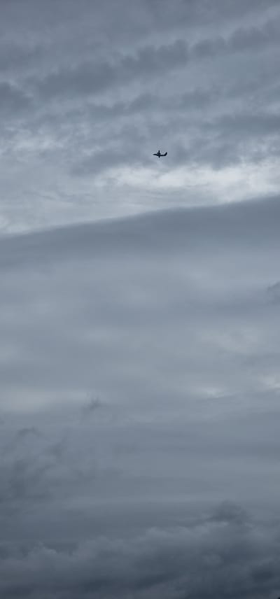 A small, dark silhouette of a twin-engine airplane is visible against a cloudy, overcast sky. The plane is positioned in the upper center of the frame. The clouds appear thick and layered, with a muted gray color palette, suggesting a dull or potentially rainy day. There is no discernible landscape or city visible, as the image is solely focused on the sky and the aircraft.